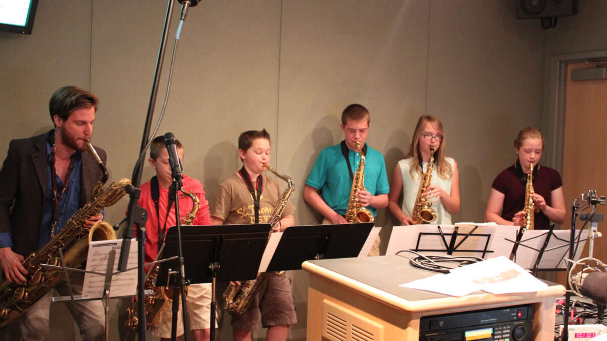 An adult playing a baritone saxophone, two youth playing tenor saxophones, two youth playing alto saxophones, and a youth playing a soprano saxophone, all looking at sheet music on music stands in a radio studio