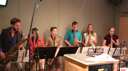 An adult playing a baritone saxophone, two youth playing tenor saxophones, two youth playing alto saxophones, and a youth playing a soprano saxophone, all looking at sheet music on music stands in a radio studio