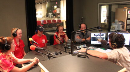 Four teens and an adult wearing headphones speaking into microphones being interviewed by an adult wearing headphones sitting behind a microphone in a radio studio