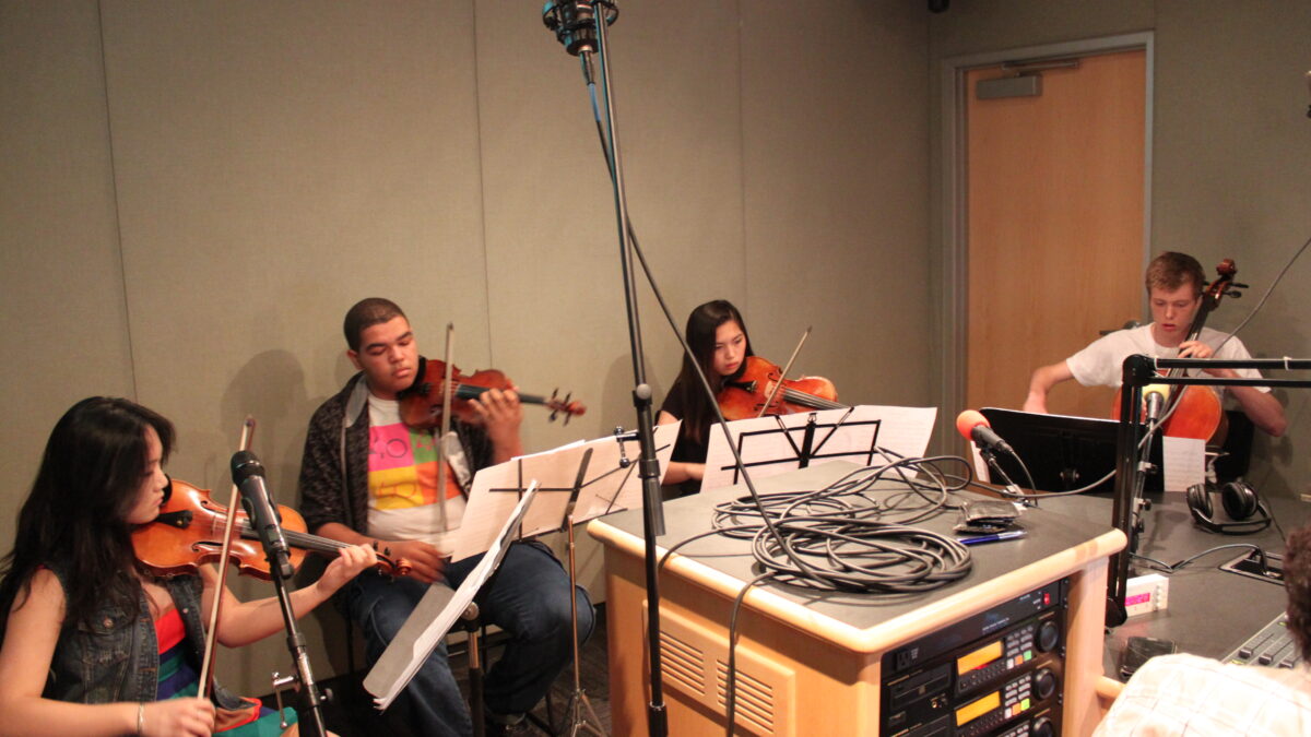 Two teens playing violin, one teen playing viola, and one teen playing cello, all looking at sheet music on music stands in a radio studio
