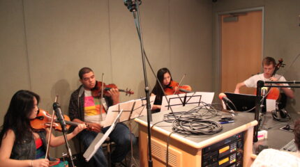 Two teens playing violin, one teen playing viola, and one teen playing cello, all looking at sheet music on music stands in a radio studio