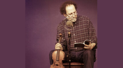 An adult looking down at a book while sitting behind a microphone and holding a violin in front of a purple background