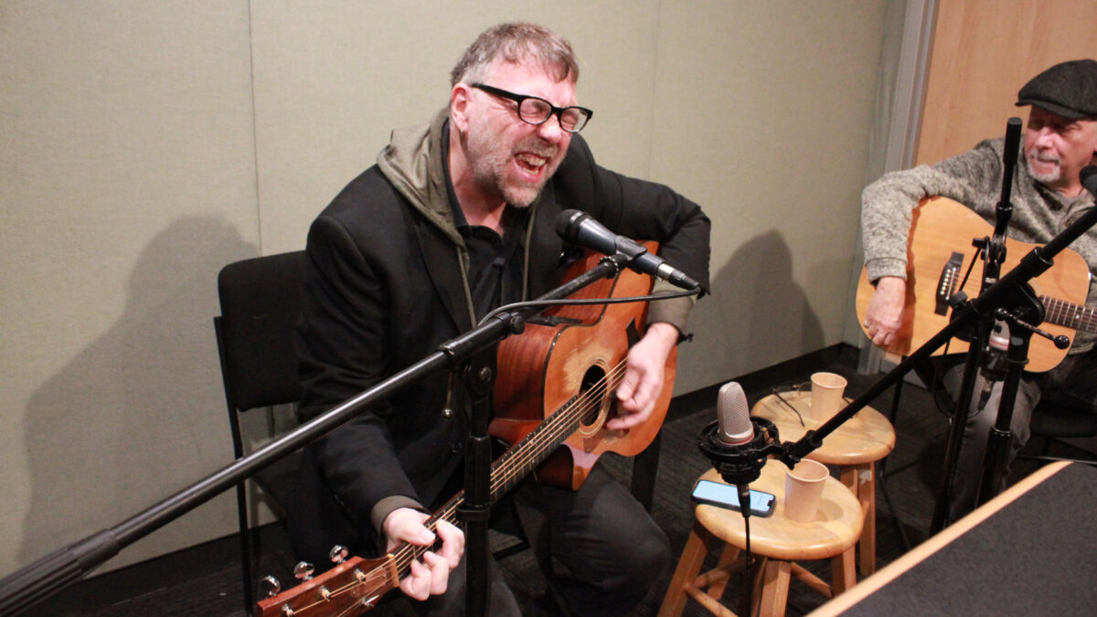 Mark-Dignam_Bill-Toms-1 An adult playing an acoustic guitar singing into a microphone and an adult sitting behind a microphone holding an acoustic guitar in a radio studio