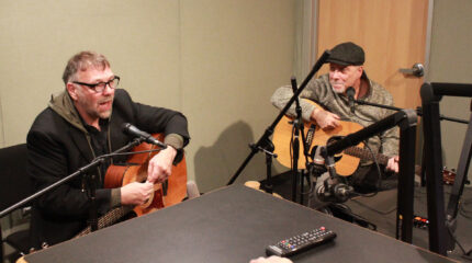 Two adults speaking into microphones and holding acoustic guitars in a radio studio
