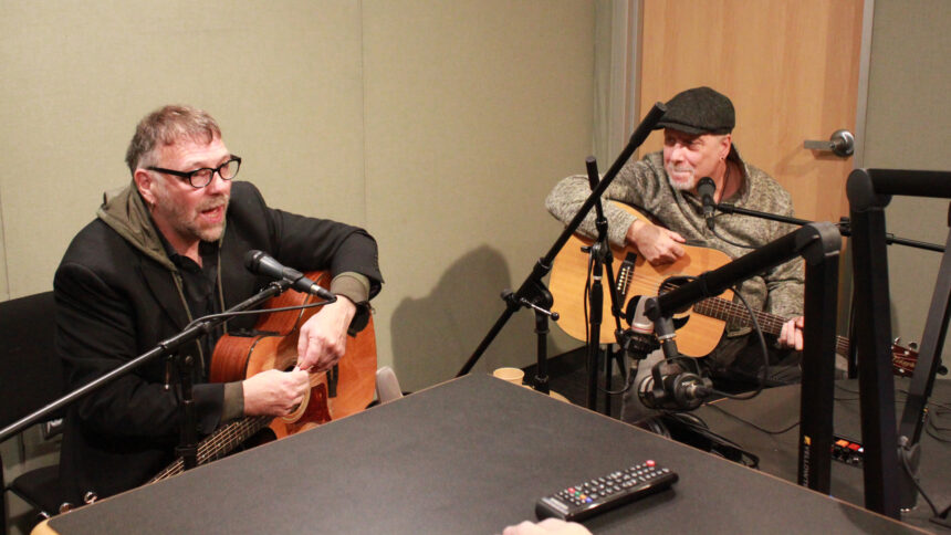 Two adults speaking into microphones and holding acoustic guitars in a radio studio