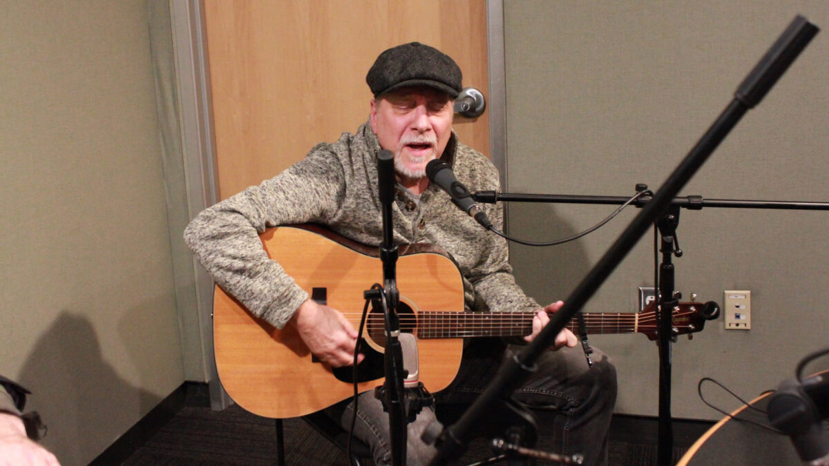 Mark-Dignam_Bill-Toms-3 An adult playing acoustic guitar and singing into a microphone in a radio studio