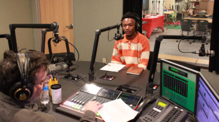 A teen wearing headphones speaking into a microphone being interviewed by an adult wearing headphones sitting behind a microphone and interacting with a mixer in a radio studio