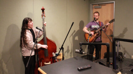 An adult playing upright bass singing into a microphone and an adult playing acoustic guitar singing into a microphone in a radio studio