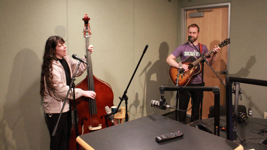 An adult playing upright bass singing into a microphone and an adult playing acoustic guitar singing into a microphone in a radio studio