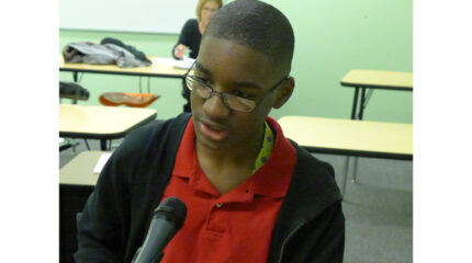 A youth speaking into a microphone standing in a green classroom