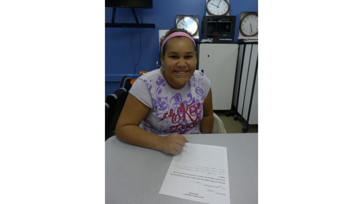 A youth smiling while sitting behind a piece of paper on a gray desk