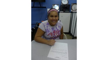 A youth smiling while sitting behind a piece of paper on a gray desk