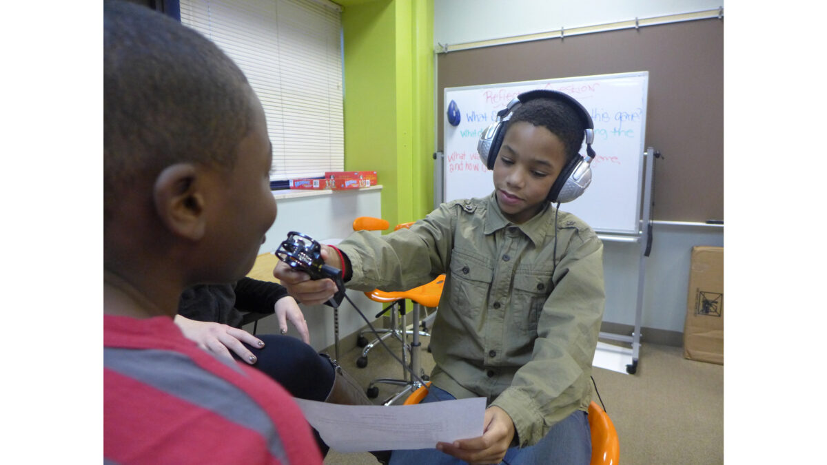 A youth wearing headphones holding a piece of paper and a portable recorder up to a youth in a classroom