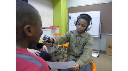 A youth wearing headphones holding a piece of paper and a portable recorder up to a youth in a classroom