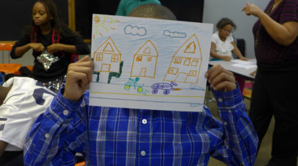 A youth hiding their face behind a drawing of a bike and a car driving in front of three houses with a sky-blue sky and a yellow sun in the background