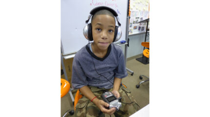 A youth wearing headphones holding a portable recorder sitting in front of a whiteboard in a classroom