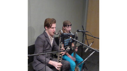 An adult and a youth playing bass clarinets looking at sheet music on a music stand in a radio studio