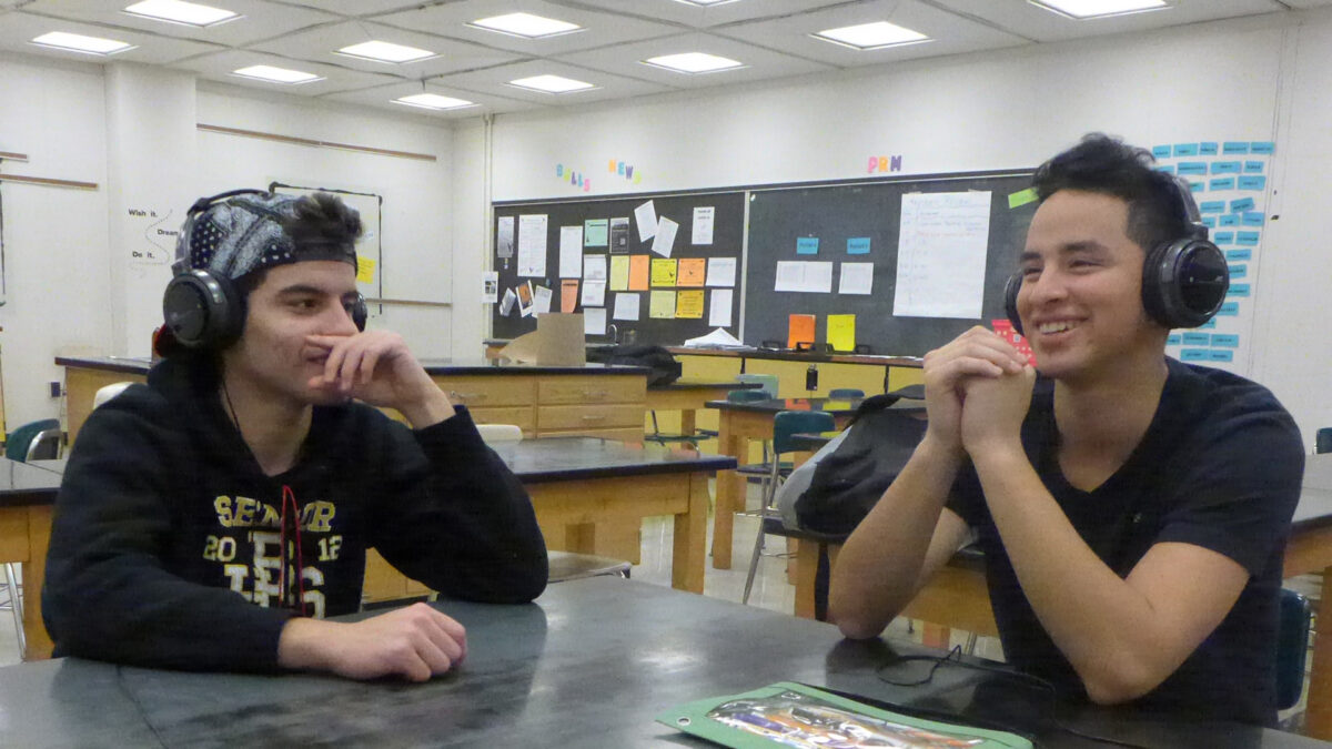 Two teens wearing headphones sitting at a black desk in a classroom
