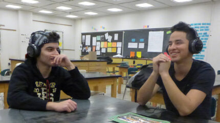 Two teens wearing headphones sitting at a black desk in a classroom