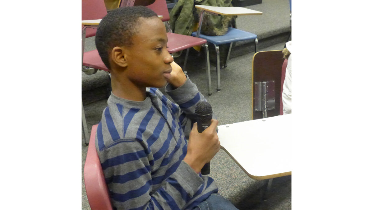 A youth holding a microphone sitting at a classroom desk