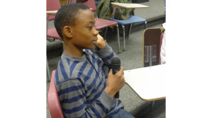 A youth holding a microphone sitting at a classroom desk