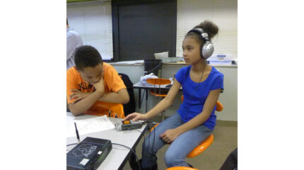 A youth looking down at a portable recorder and a youth wearing headphones, both sitting in orange chairs in a classroom
