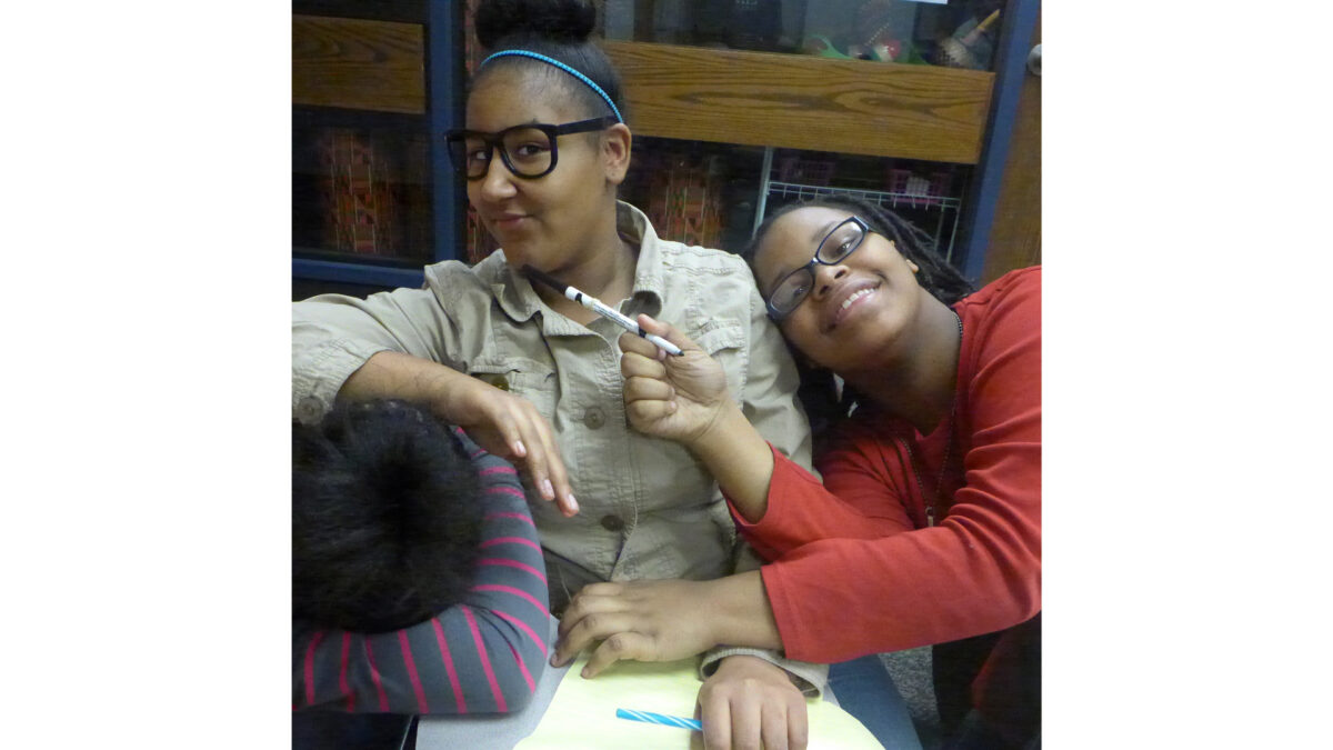 Two youth smiling and sitting while holding markers in a classroom