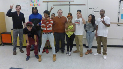 Eight teens and two adults smiling and posing in a classroom