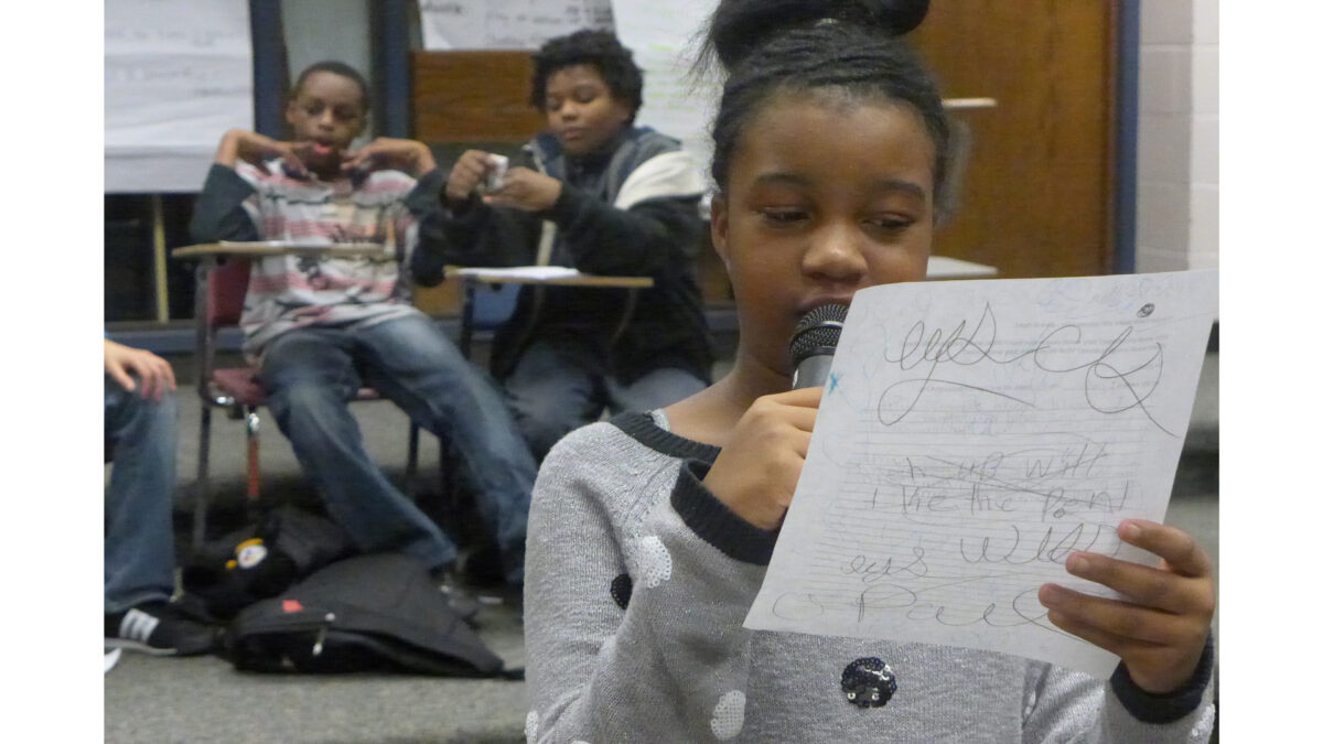 A youth holding a piece of paper and speaking into a microphone as two youth sit in classroom desks in the background