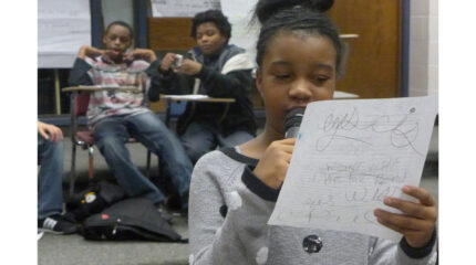 A youth holding a piece of paper and speaking into a microphone as two youth sit in classroom desks in the background