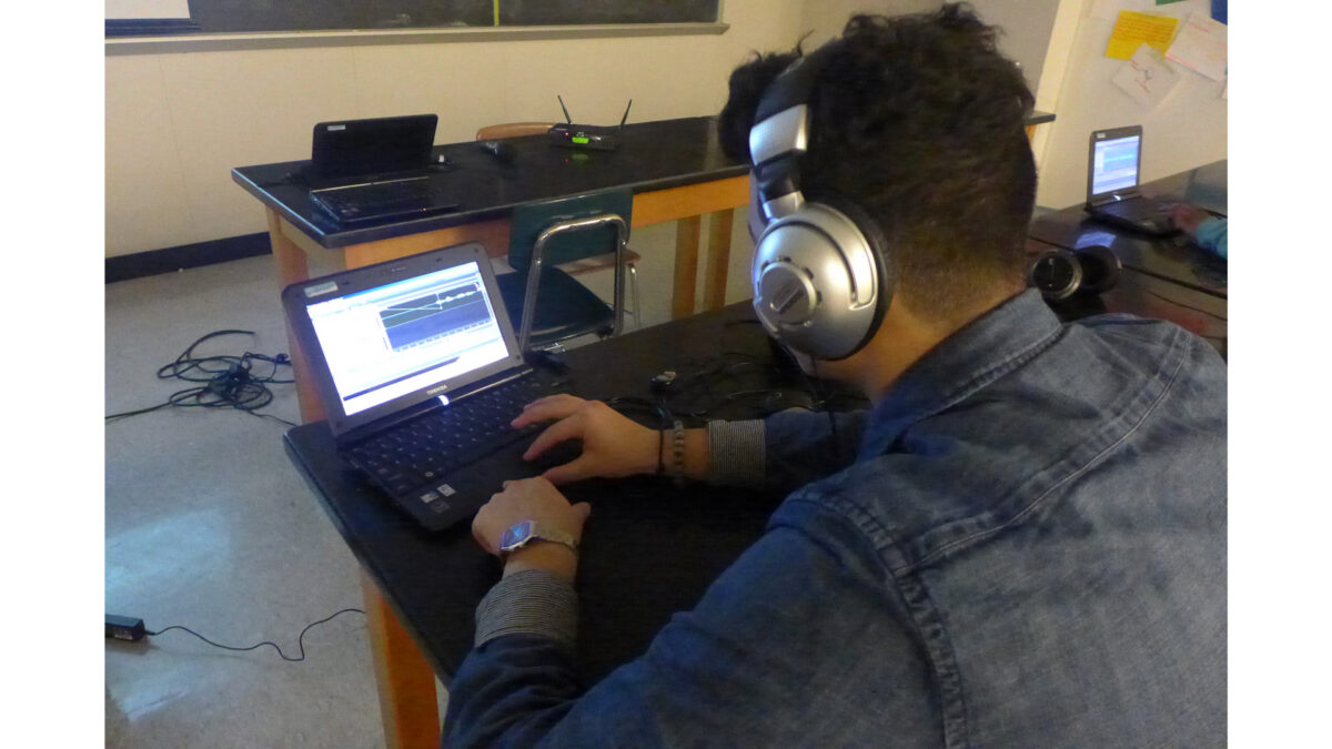 A teen wearing headphones looking at audio editing software on a laptop computer in a classroom