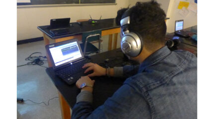 A teen wearing headphones looking at audio editing software on a laptop computer in a classroom