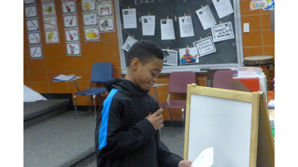A youth looking down at a piece of paper and holding and speaking into a microphone in a classroom