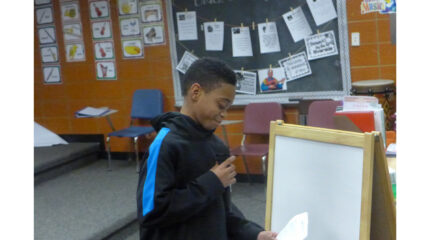 A youth looking down at a piece of paper and holding and speaking into a microphone in a classroom