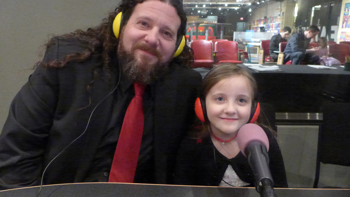 An adult and a youth wearing headphones sitting behind microphones in a radio studio