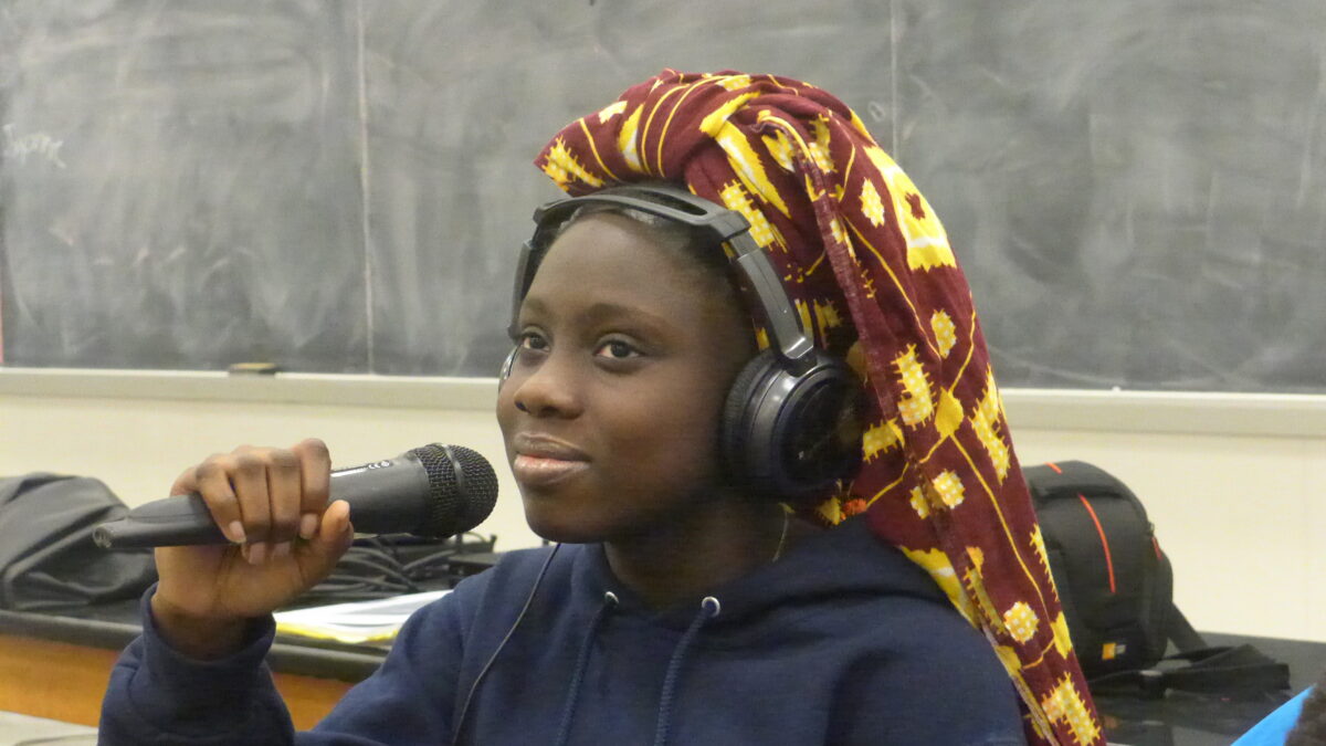 A teen wearing headphones and a head scarf holding a microphone in a classroom