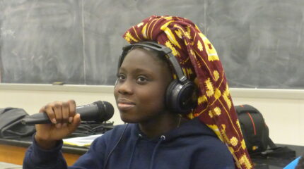 A teen wearing headphones and a head scarf holding a microphone in a classroom