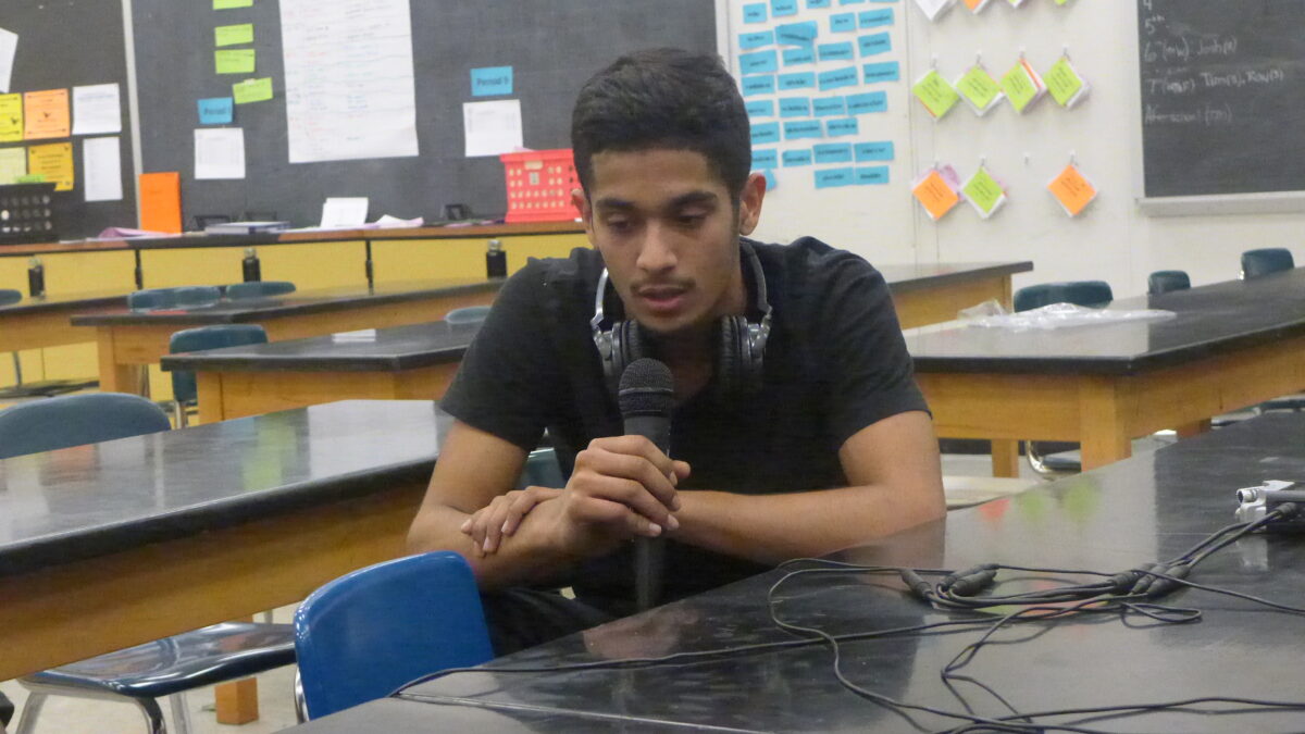 A teen wearing headphones around their neck speaking into a microphone in a classroom