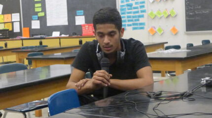 A teen wearing headphones around their neck speaking into a microphone in a classroom