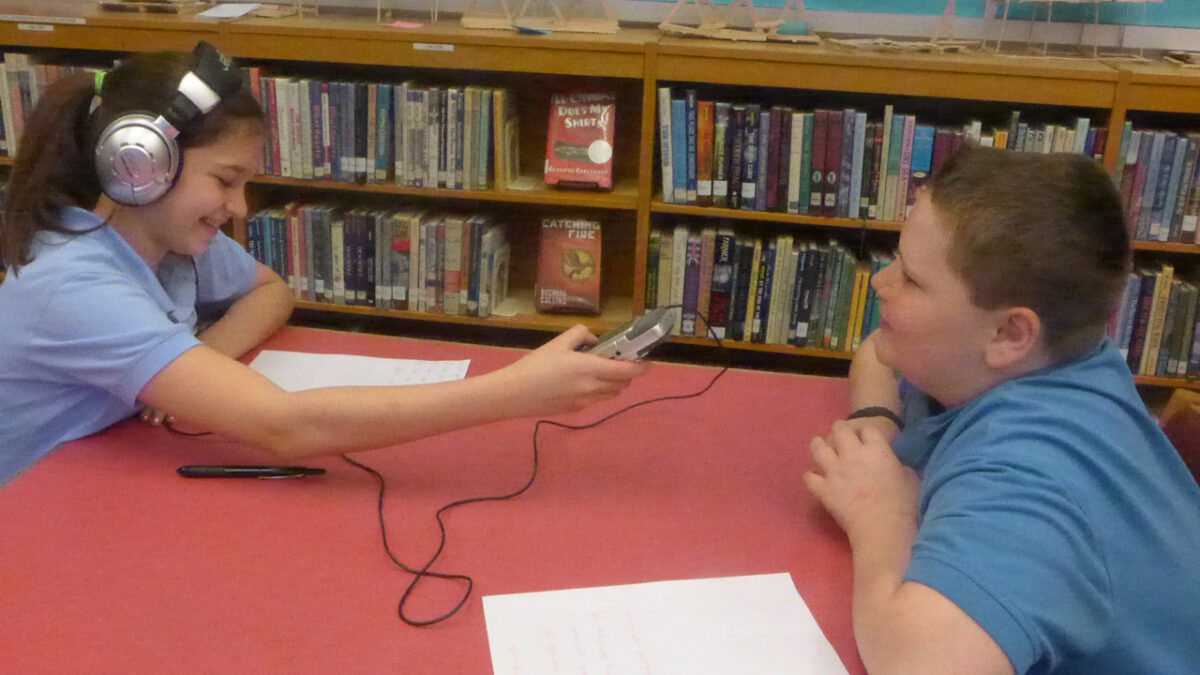 A youth wearing headphones looking down at a piece of paper and holding up a portable recorder interviewing another youth in a library