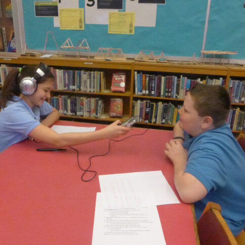 A youth wearing headphones looking down at a piece of paper and holding up a portable recorder interviewing another youth in a library