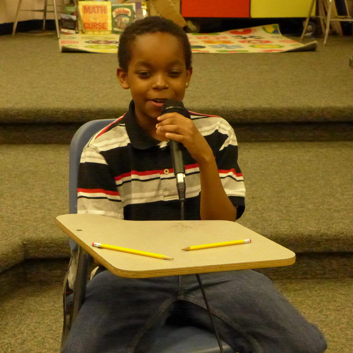 A youth sitting at a classroom desk holding and speaking into a microphone