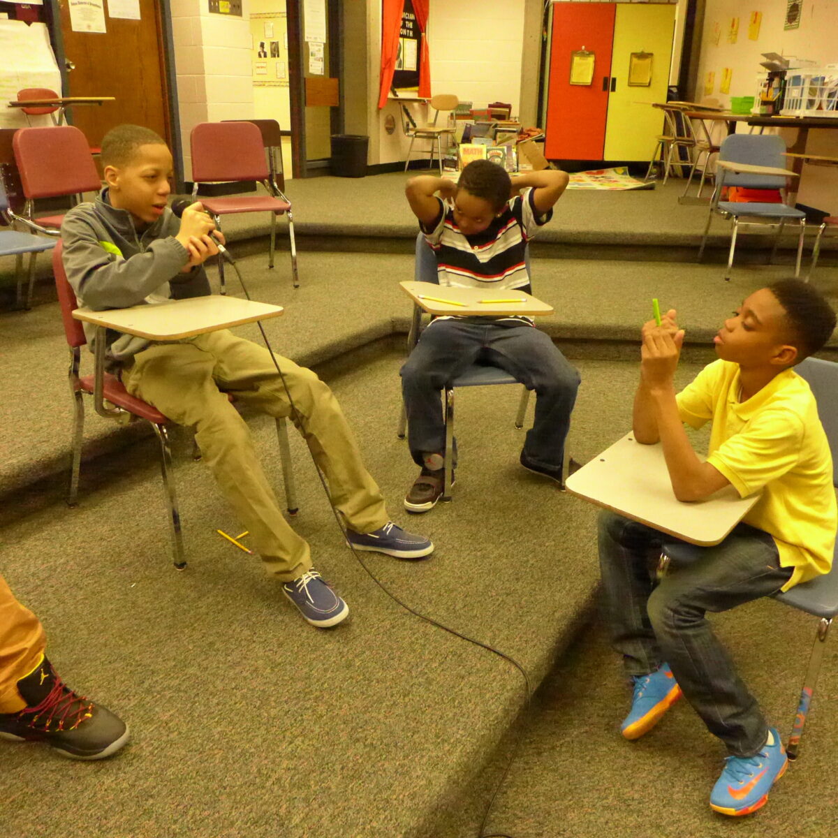 Three youth sitting at classroom desks, one holding and speaking into a microphone