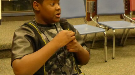 A youth sitting at a classroom desk holding and speaking into a microphone