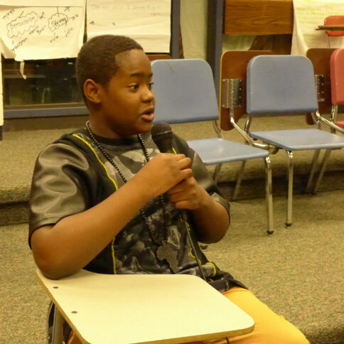 A youth sitting at a classroom desk holding and speaking into a microphone