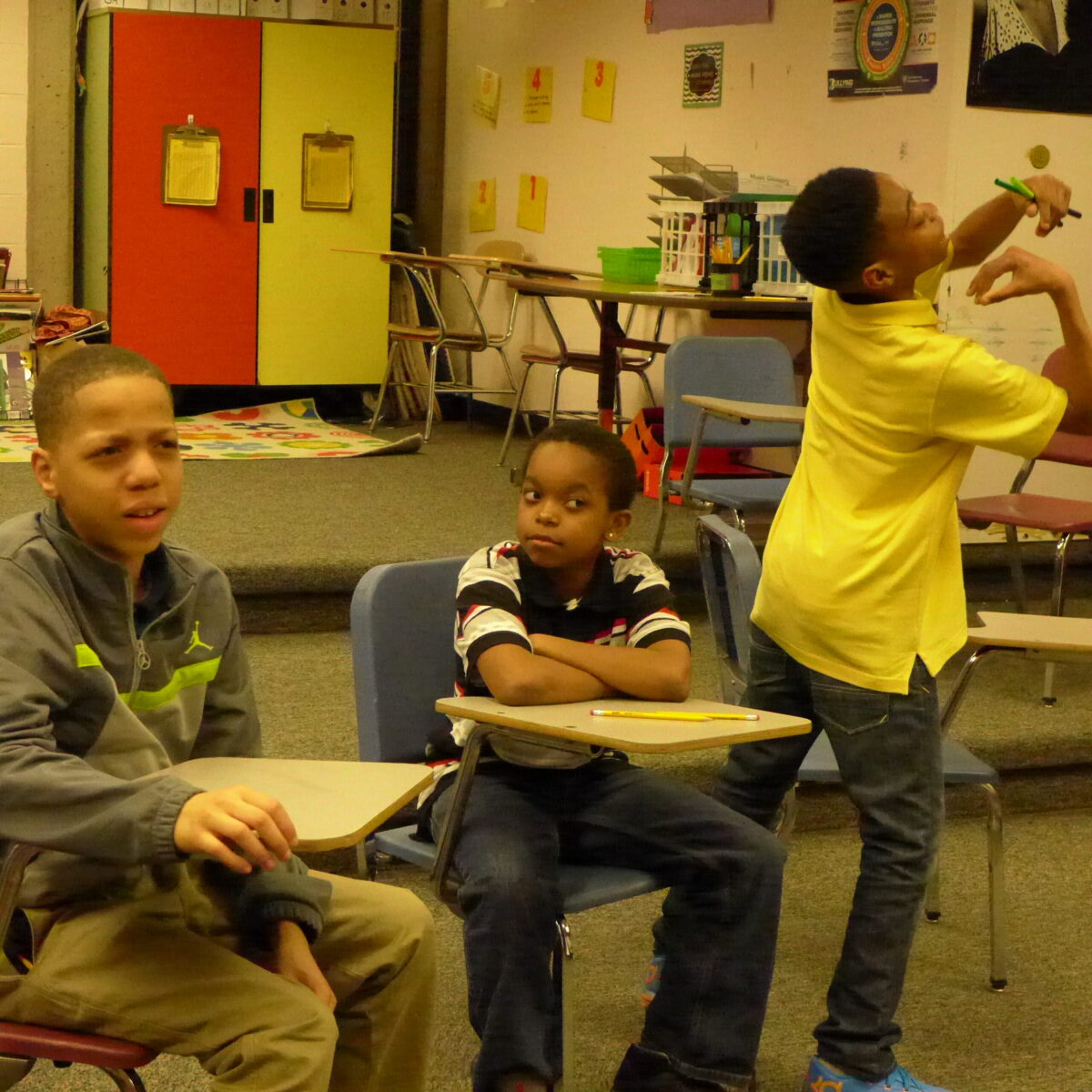 Three youth in a classroom, two sitting while making inquisitive faces, the other posing while looking to the right