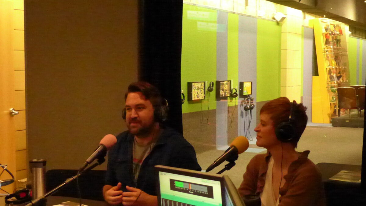Two adults wearing headphones speaking into microphones in a radio studio