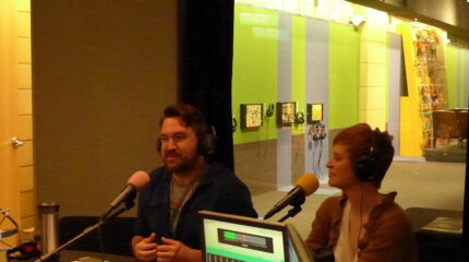 Two adults wearing headphones speaking into microphones in a radio studio