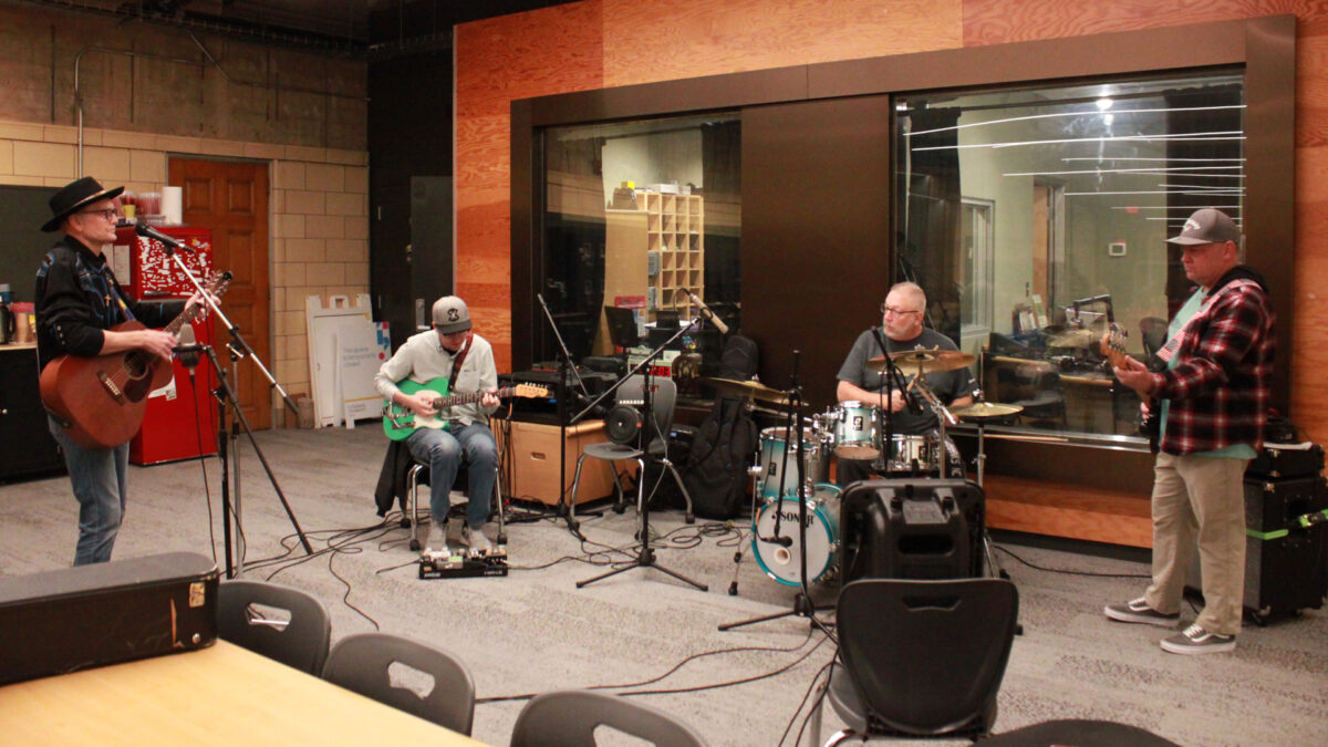 Slim-Forsythe-and-the-Boss-Stingrays-1 An adult playing acoustic guitar, an adult playing electric guitar, an adult playing a drum set, and an adult playing electric bass