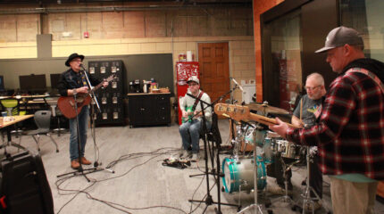 An adult playing acoustic guitar and singing into a microphone, an adult playing electric guitar, an adult playing drum set, and an adult playing electric bass